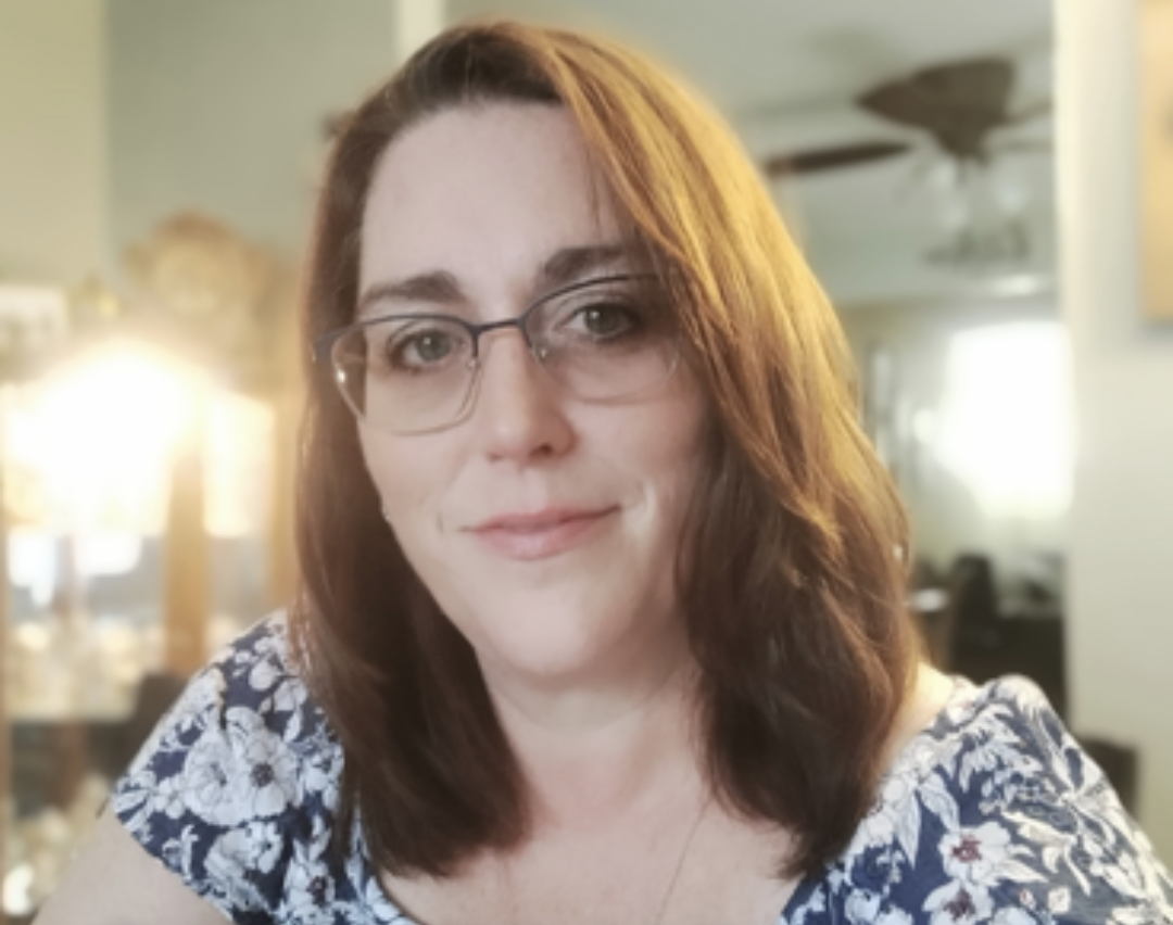 Woman with shoulder-length brown hair and glasses wearing a blue and white floral top indoors.
