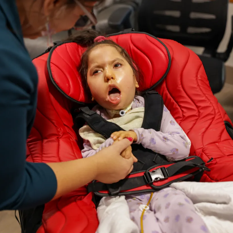 A young child with medical needs sits in a supportive red adaptive chair, wearing a tracheostomy tube and patterned pajamas, while an adult gently holds the child’s hand and offers support. The child looks upward with an open mouth expression, and the scene takes place in an indoor care setting.