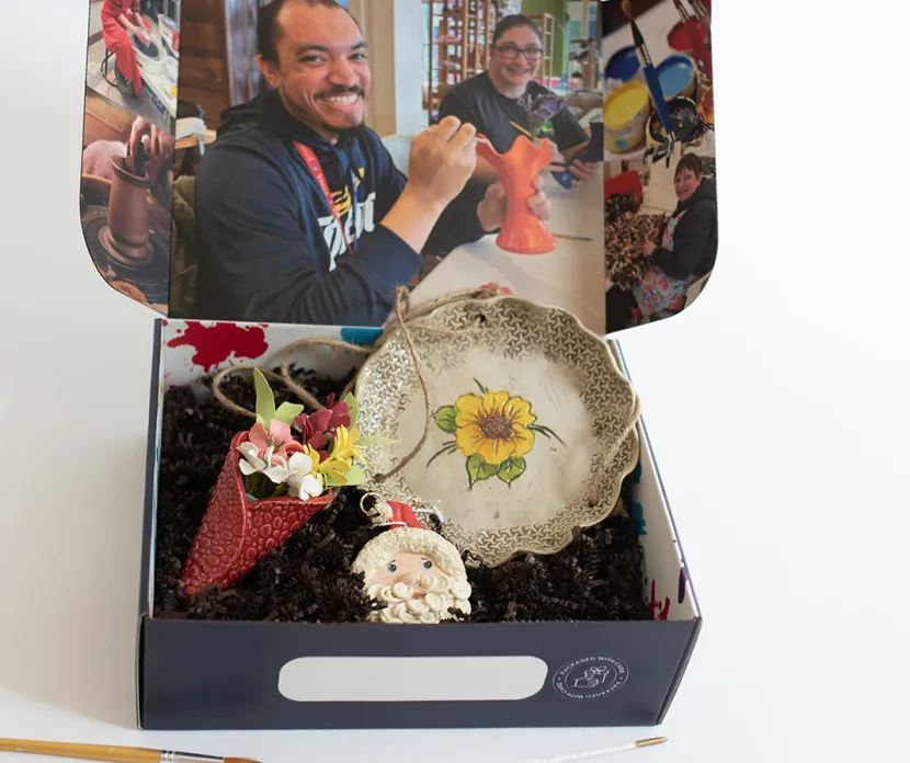 Art gift box featuring handcrafted pottery, including a sunflower-themed ceramic plate, a Santa ornament, and a colorful paper flower arrangement. The inside of the box is decorated with images of smiling artisans working on their creations. Paintbrushes are scattered around, emphasizing the handmade artistry of the items.