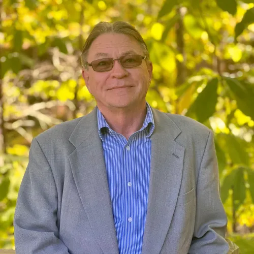 "A man wearing glasses, a light gray blazer, and a blue striped shirt stands outdoors with a calm expression. He is posed in front of a background filled with lush green leaves, giving the image a natural and serene feel."