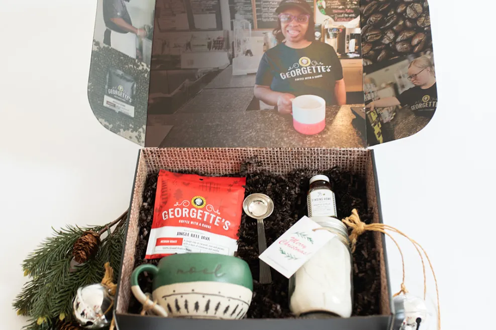 Alt text: "The Christmas Morning gift box from Georgette’s, featuring a festive collection including a bag of Jingle Bell Java coffee, a handmade ceramic mug with 'Noel' on it, a jar of powdered drink mix, and a coffee scoop. The box is decorated with holiday greenery and bells, with photos of Georgette’s employees displayed inside the lid."