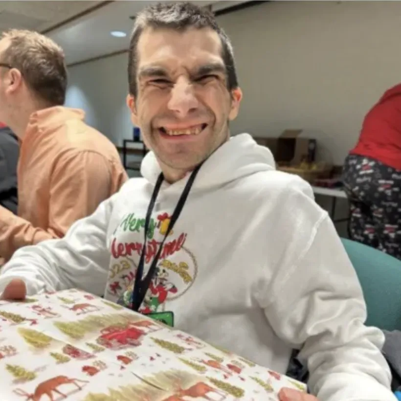 Smiling man holding a Christmas gift wrapped in festive holiday paper, wearing a white Merry Christmas hoodie at a holiday celebration.