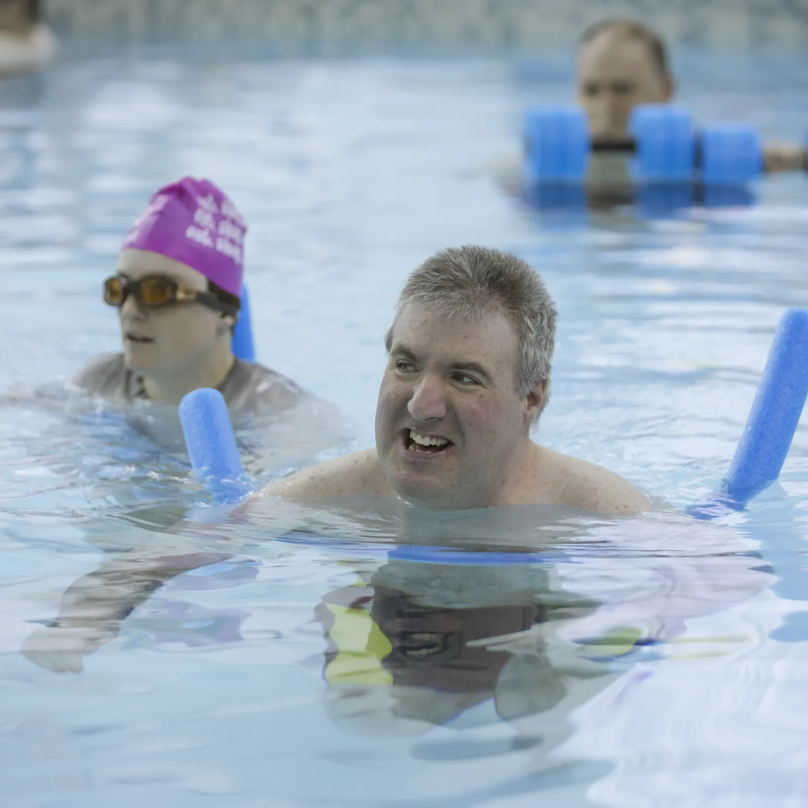 Adults with developmental disabilities participate in aquatic therapy at a community pool, using flotation devices and water weights to support health, mobility, and well-being during a therapeutic recreation session.