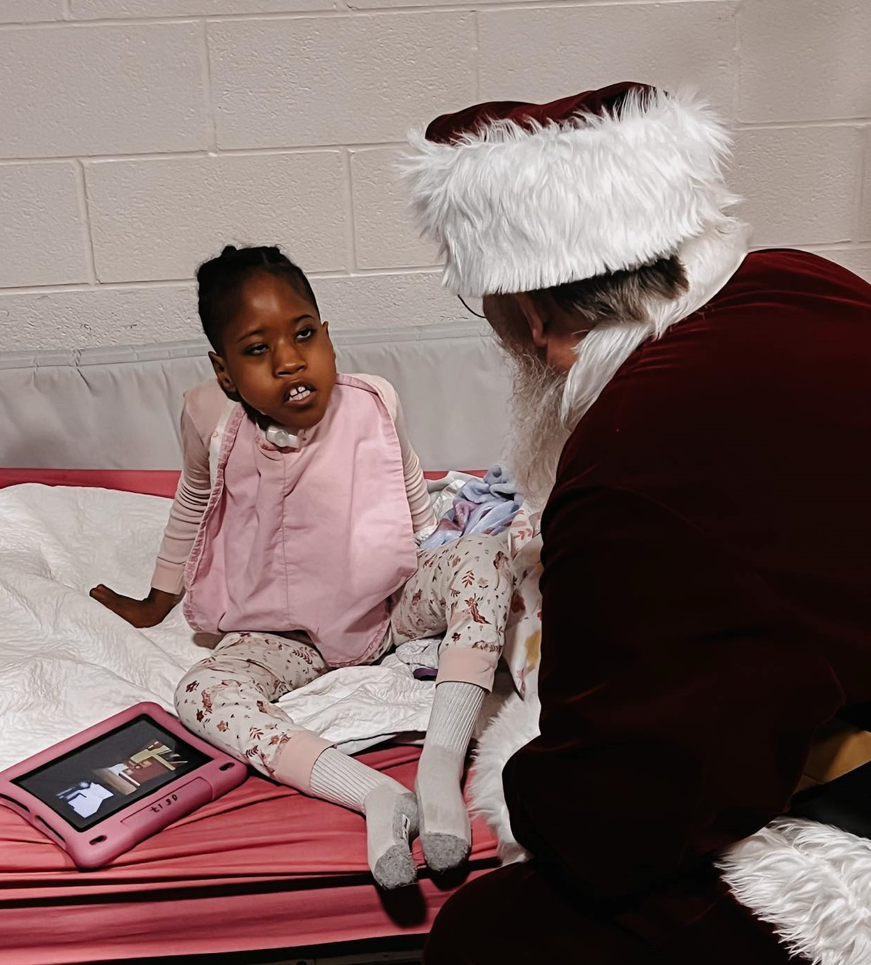 Girl on pink bed with Santa, smiling beside her tablet.