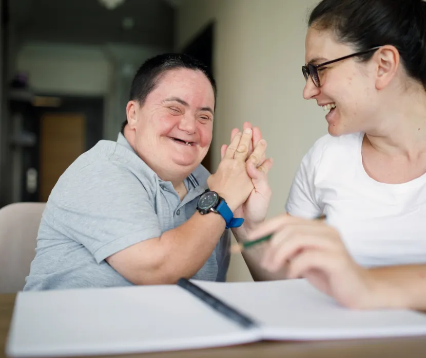 Smiling woman with Down syndrome shares a joyful moment with her caregiver at a table, celebrating connection, inclusion, and supportive disability services.