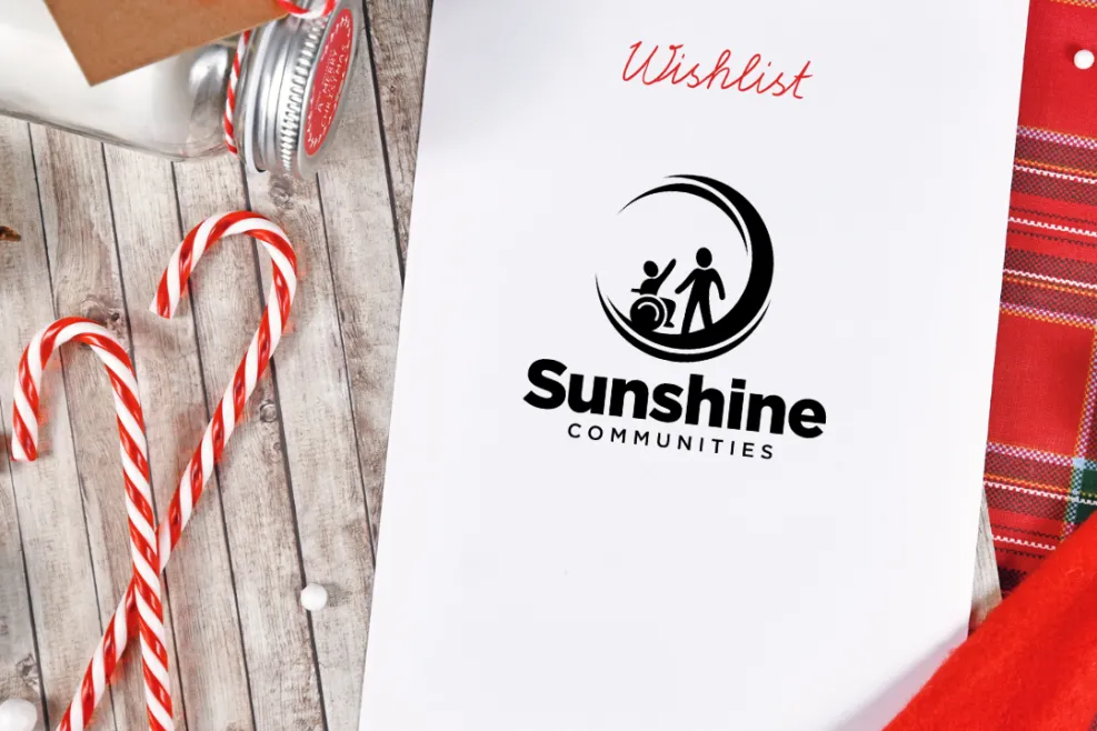 Alt Text: A holiday-themed setup featuring a "Wishlist" document with the Sunshine Communities logo on the cover, surrounded by festive items including candy canes, snowflake decorations, a jar with a red-striped bow, and a Santa hat. The background is a rustic wooden surface with a plaid fabric adding a warm holiday touch.