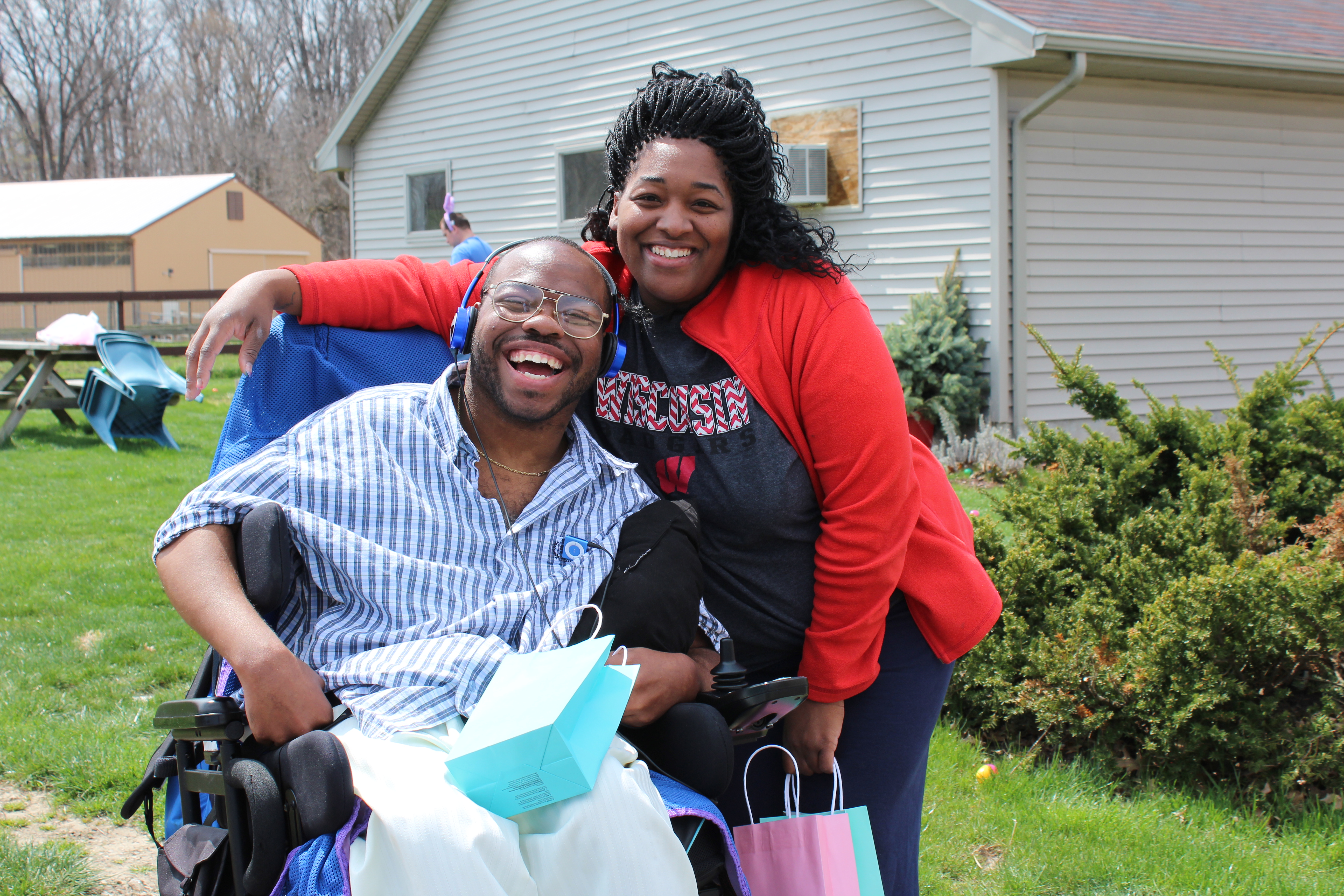 A young man in a wheelchair with glasses and blue headphones is smiling joyfully, holding a light blue gift bag. Beside him, a staff member wearing a red jacket and a Wisconsin Badgers t-shirt leans in with a warm smile. They are outdoors on a grassy lawn with picnic tables, a beige house, and a barn in the background. The scene conveys happiness, support, and community.