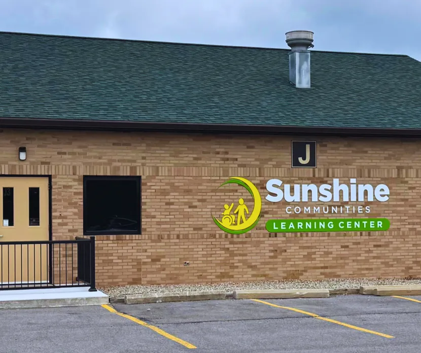 Exterior view of Sunshine Communities Learning Center building, featuring accessible entrance, designated parking, and bold signage with the organization's logo.
