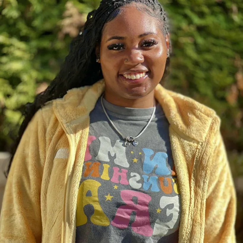 A young woman stands outdoors in bright sunlight, smiling warmly at the camera.
