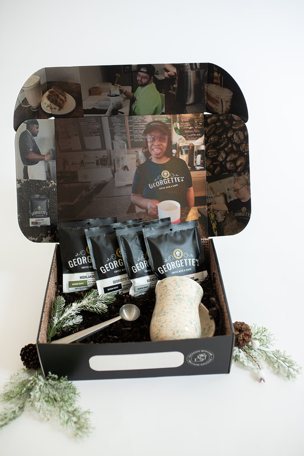 Holiday gift box from Georgette's featuring four bags of medium roast coffee, a ceramic mug, and a metal scoop, surrounded by festive pine sprigs. The inside of the box showcases photos of Georgette's employees, emphasizing the café's inclusive mission."