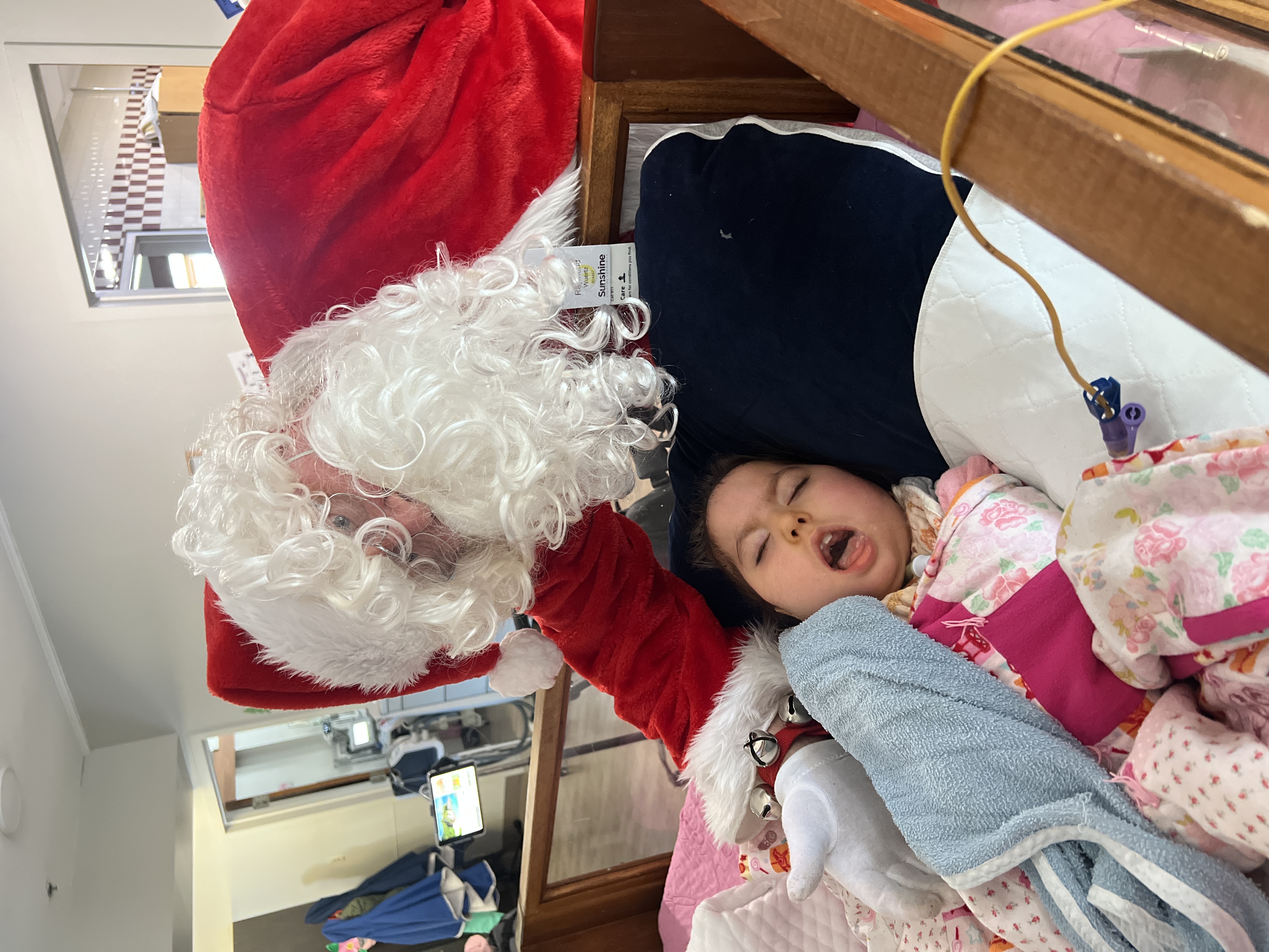 Santa leans over a child resting in bed during a holiday visit at Sunshine, sharing a warm and joyful Christmas moment.