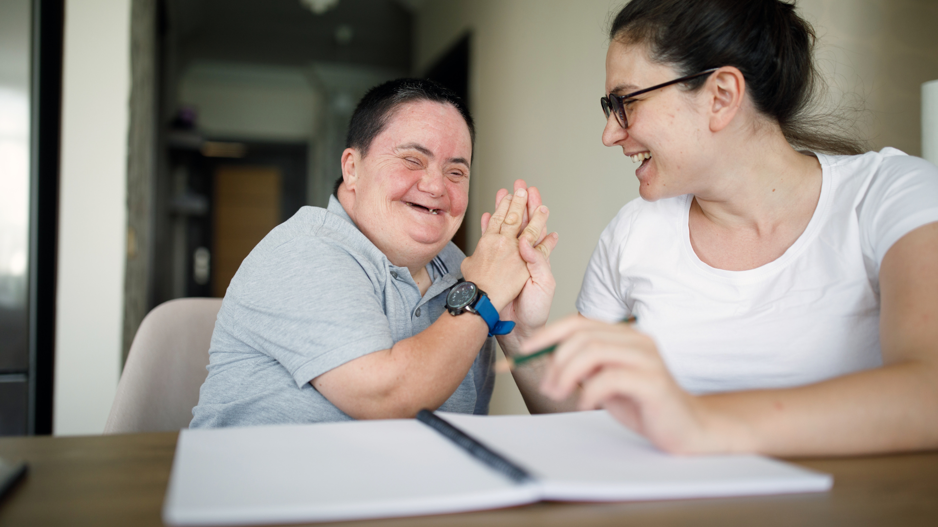 Smiling woman with Down syndrome shares a joyful moment with her caregiver at a table, celebrating connection, inclusion, and supportive disability services.