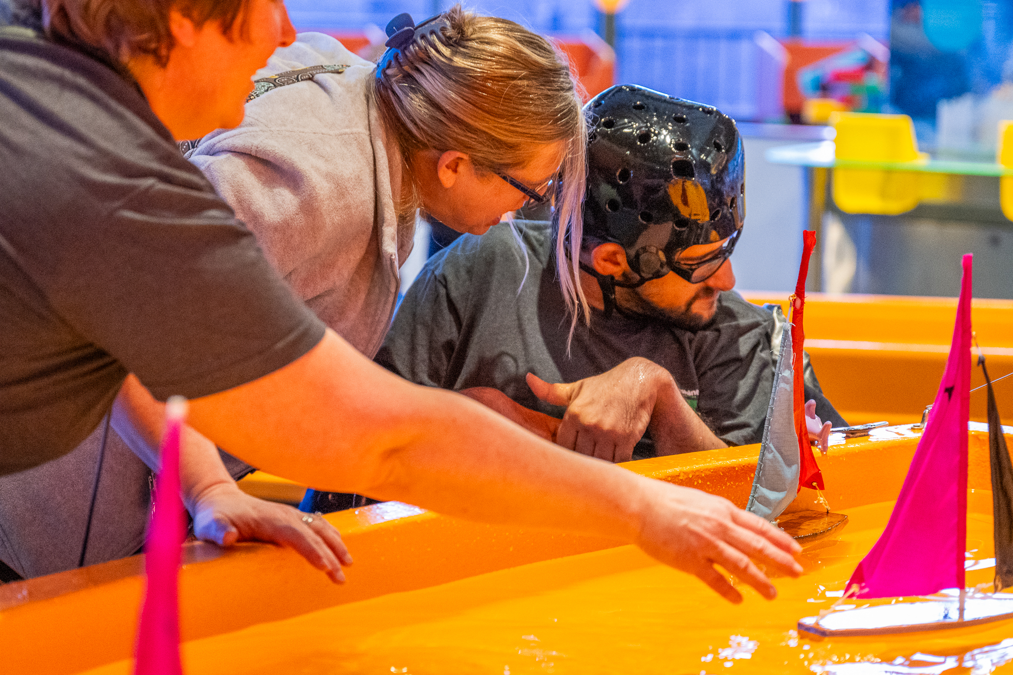 A sensory-friendly activity where a man wearing a protective helmet interacts with a water table featuring small colorful sailboats. Two caregivers assist him, one guiding his hand and the other offering support nearby. The environment is brightly lit and welcoming.