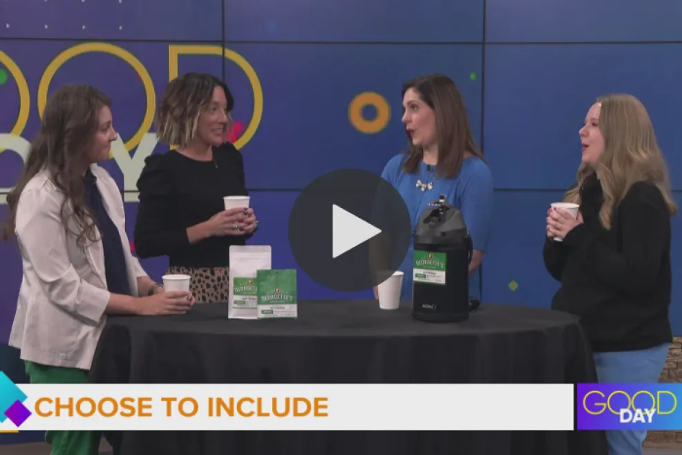 Four women stand around a small round table on a TV studio set during a “Good Day” segment labeled “Choose to Include.” They are holding white coffee cups and talking. On the table are two green-and-white Georgette’s coffee bags and a large black airpot dispenser. A blue studio backdrop with colorful circular graphics appears behind them, and a “Good Day” logo with the time (9:24) is visible in the bottom right corne