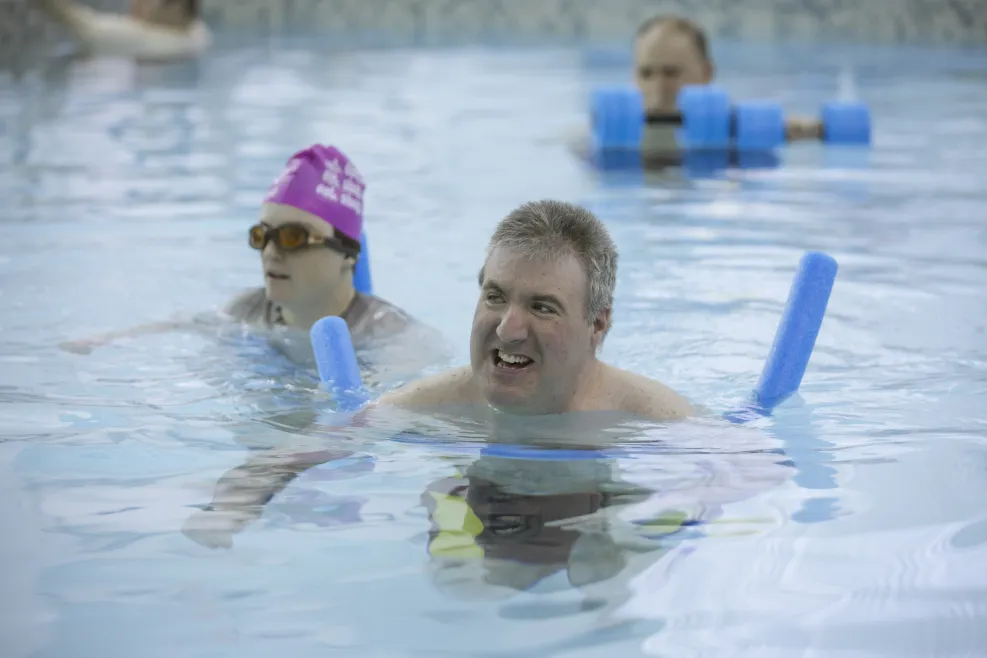 Two adults with developmental disabilities participate in an adaptive aquatic therapy session in a heated indoor pool. One swimmer wears a pink swim cap and goggles, while the other smiles, supported by a blue pool noodle. A third individual in the background uses aqua dumbbells for resistance training. This inclusive, therapeutic recreation program promotes wellness, mobility, and joy for individuals of all abilities at Sunshine Communities.