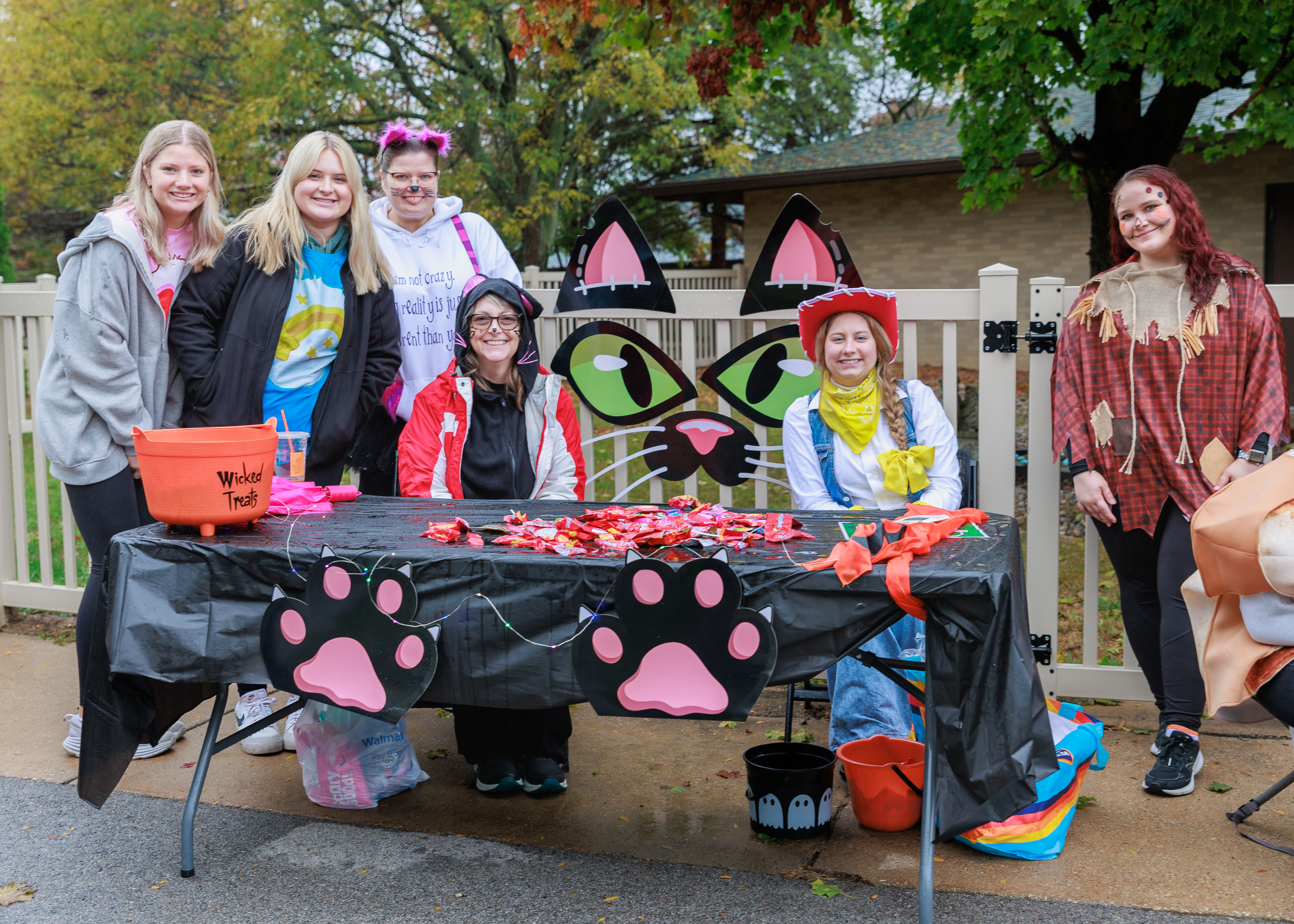 Six costumed staff members pose behind a cat-themed trick-or-treat table filled with candy during a Halloween celebration at Sunshine.