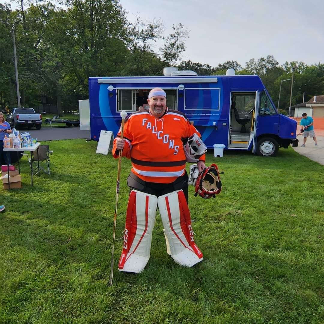 Rob Gasser dressed as a hockey goalie in a Falcons jersey standing on grass at an outdoor event with a blue truck in the background