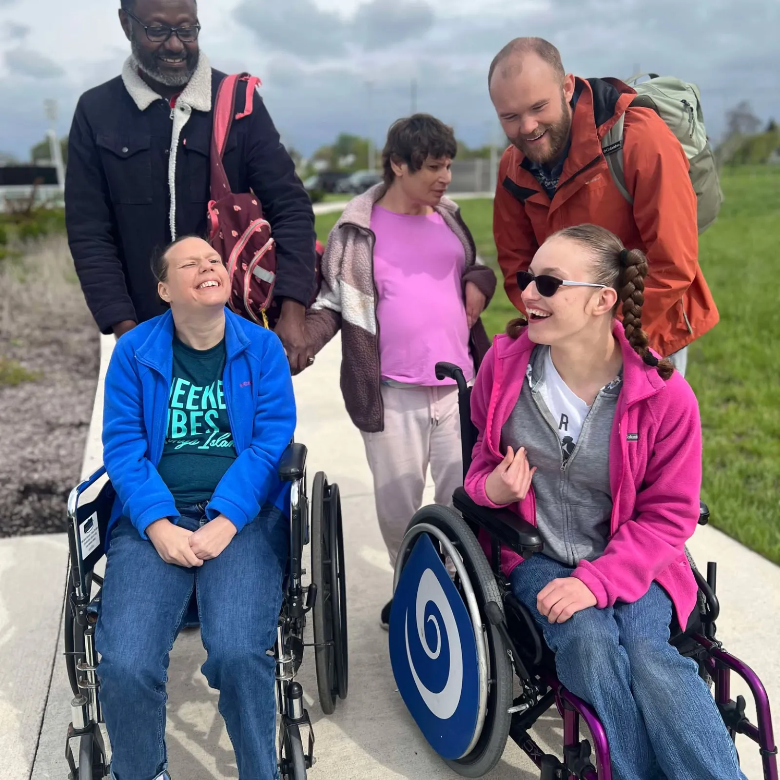 A group of five individuals smile for a photo outside. Two are in wheelchairs.