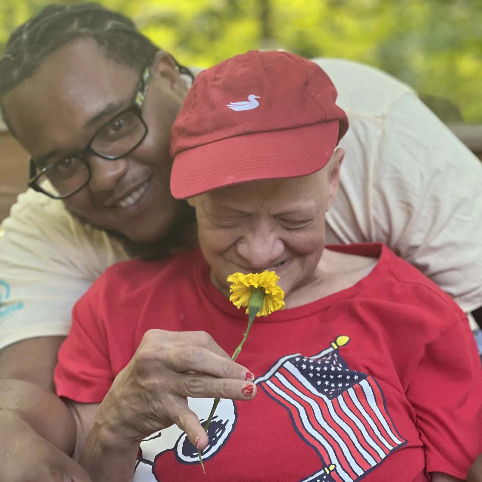 Smiling caregiver hugging an elderly woman in a red shirt as she holds and smells a yellow flower outdoors