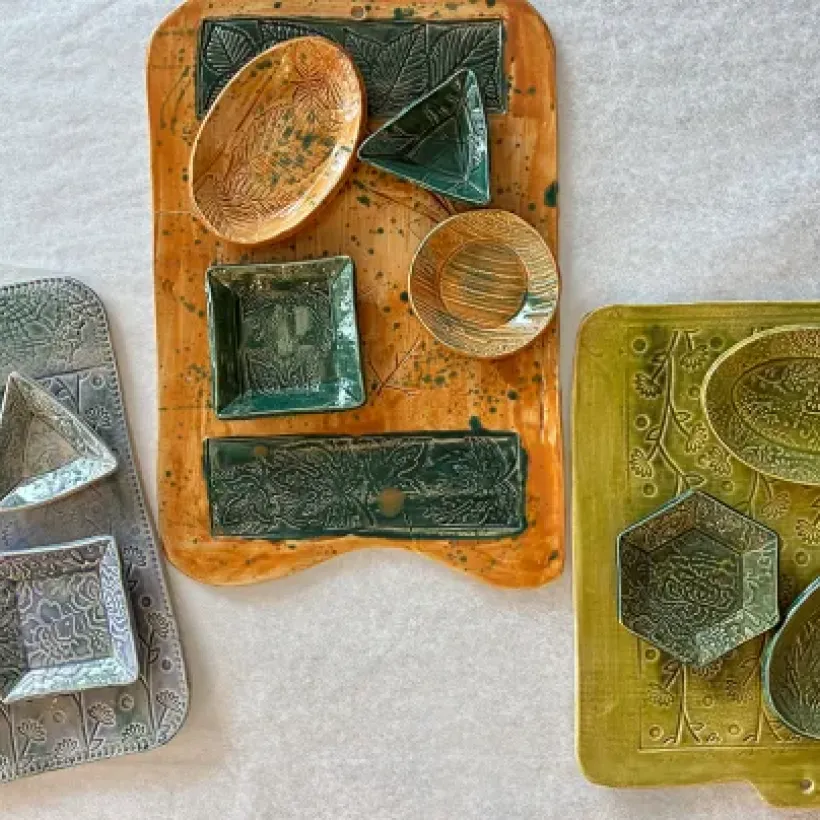 Three handcrafted ceramic charcuterie boards in green, orange, and gray glazes, featuring embossed botanical and geometric designs with matching small dishes arranged on top.