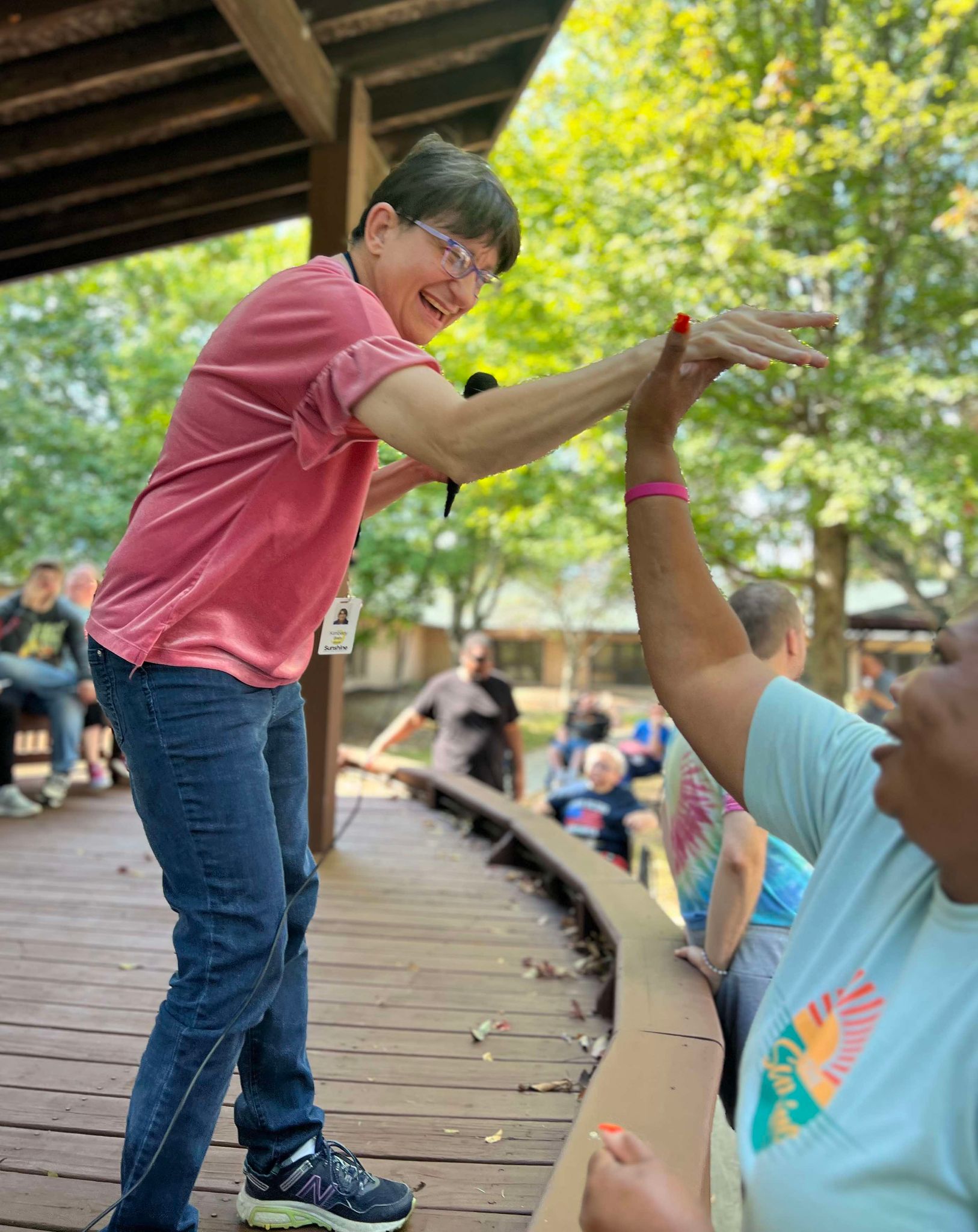 A smiling woman with short brown hair, wearing a pink shirt and jeans, gives a high-five to another person wearing a light blue Sunshine Communities T-shirt during an outdoor event under a wooden pavilion. People in the background look on, enjoying the sunny day surrounded by trees.
