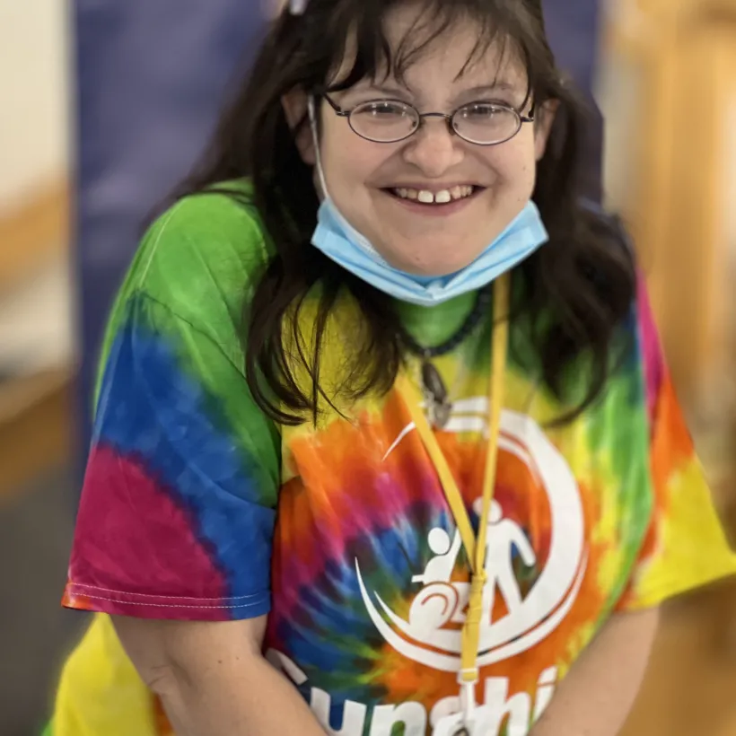 A smiling woman wearing glasses and a colorful tie-dye Sunshine Communities T-shirt sits indoors with a lanyard name badge and a face mask resting under her chin.