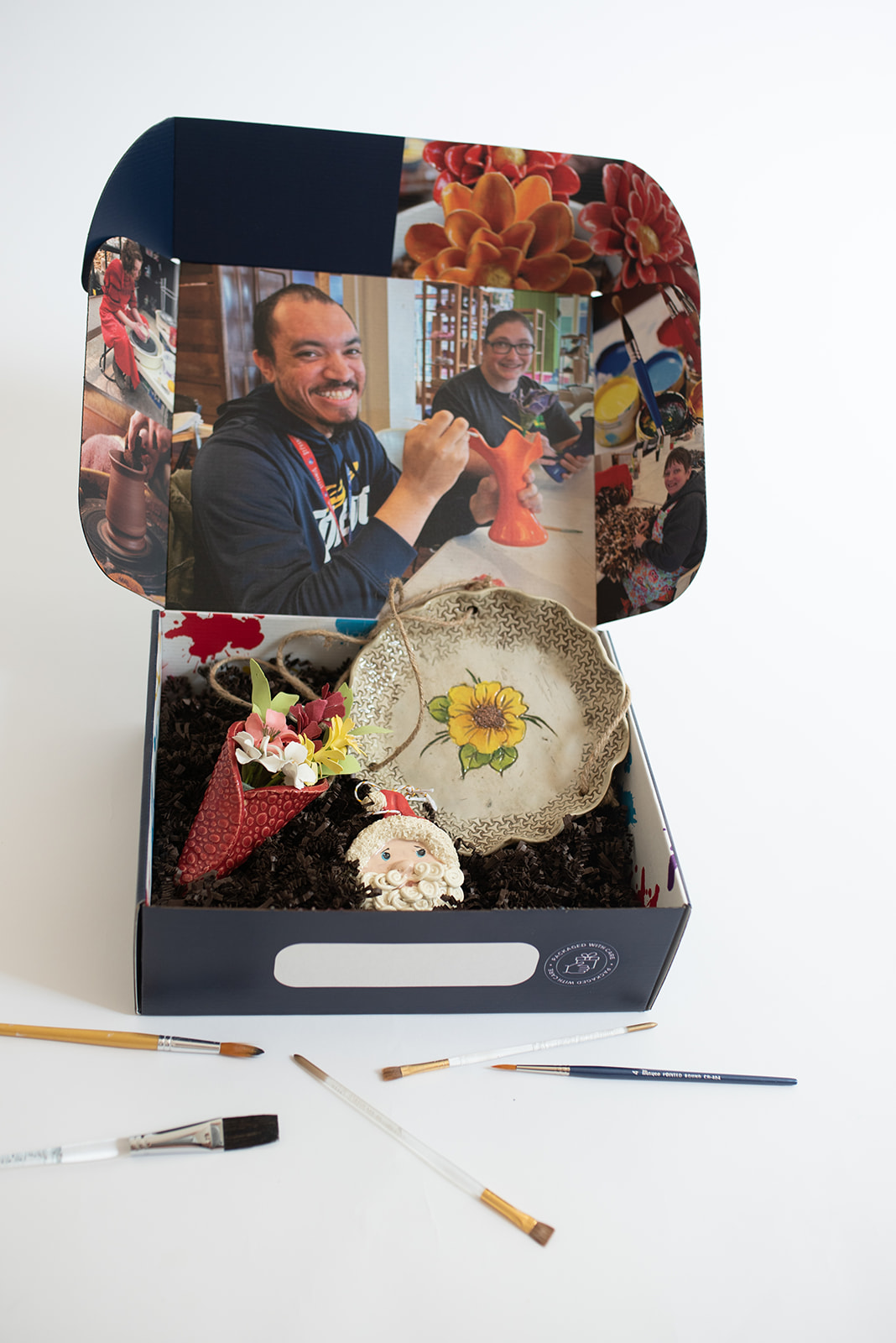 Art gift box featuring handcrafted pottery, including a sunflower-themed ceramic plate, a Santa ornament, and a colorful paper flower arrangement. The inside of the box is decorated with images of smiling artisans working on their creations. Paintbrushes are scattered around, emphasizing the handmade artistry of the items.