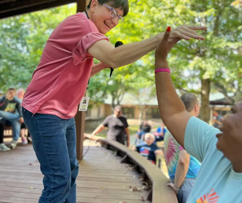 A smiling woman with short brown hair, wearing a pink shirt and jeans, gives a high-five to another person wearing a light blue Sunshine Communities T-shirt during an outdoor event under a wooden pavilion. People in the background look on, enjoying the sunny day surrounded by trees.