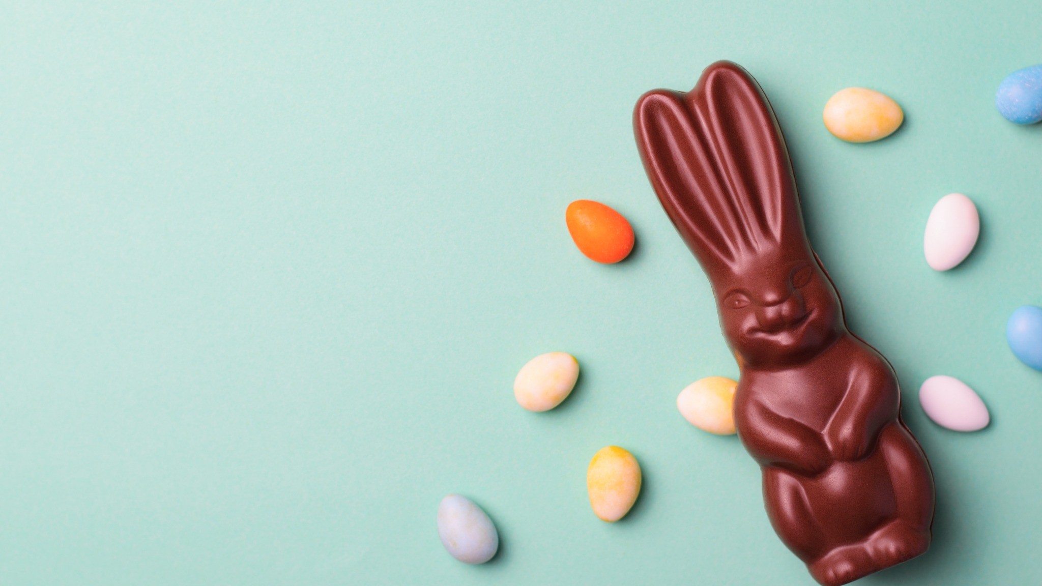 "A chocolate bunny with a glossy finish lies on a pastel green background, surrounded by colorful candy eggs in shades of orange, blue, white, and yellow."