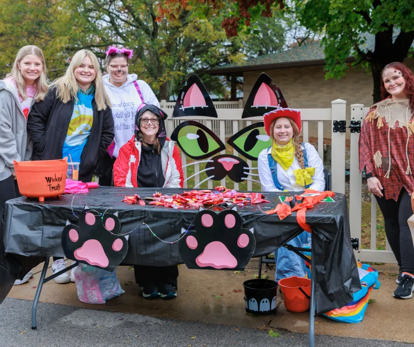 Six costumed staff members pose behind a cat-themed trick-or-treat table filled with candy during a Halloween celebration at Sunshine.