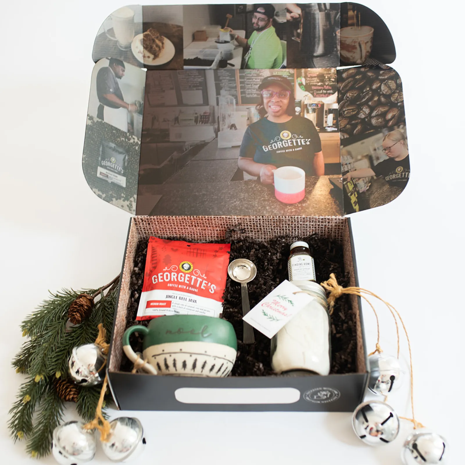 Alt text: "The Christmas Morning gift box from Georgette’s, featuring a festive collection including a bag of Jingle Bell Java coffee, a handmade ceramic mug with 'Noel' on it, a jar of powdered drink mix, and a coffee scoop. The box is decorated with holiday greenery and bells, with photos of Georgette’s employees displayed inside the lid."