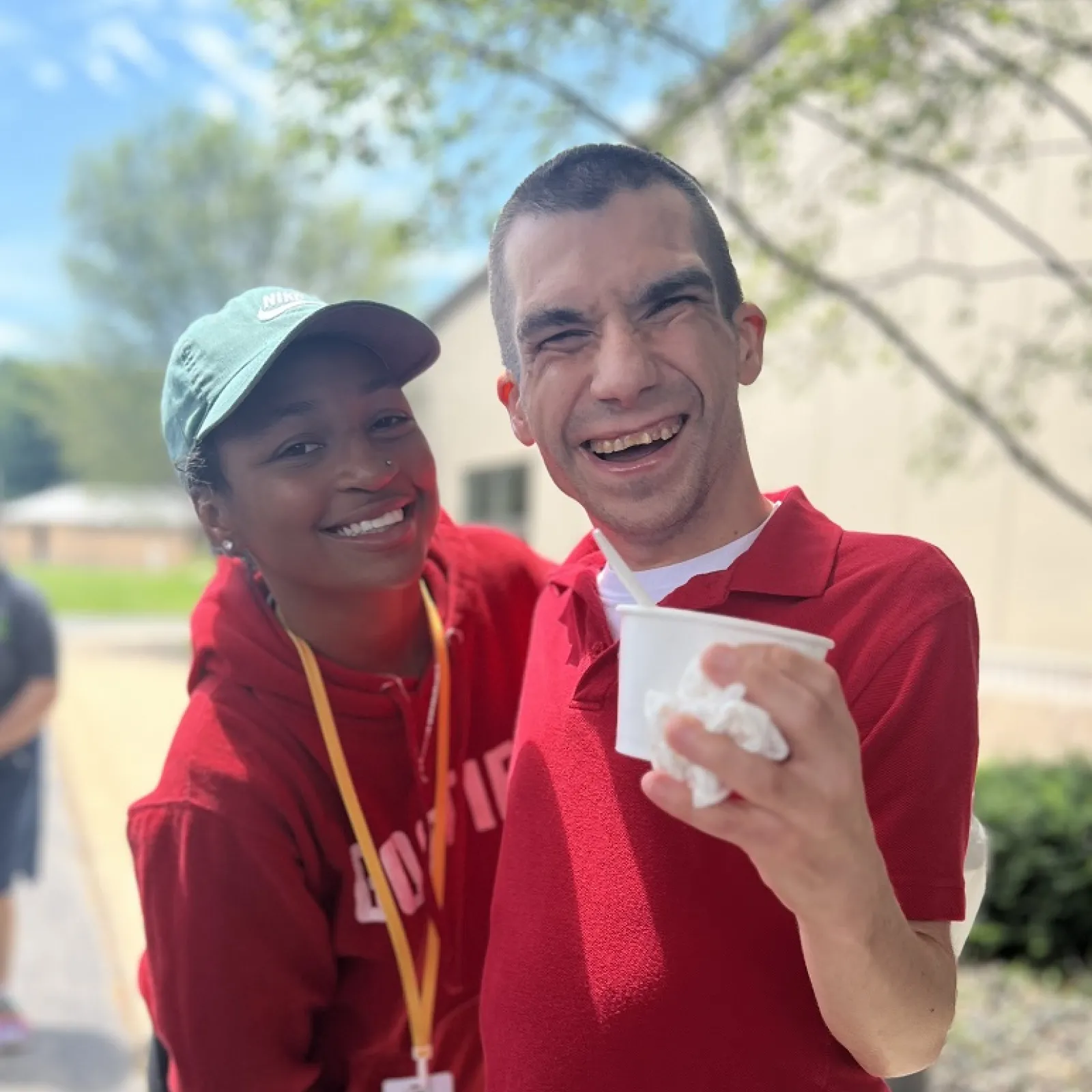 "A smiling young man in a red shirt holds a cup of ice cream while posing with a woman in a red hoodie and green cap. They are outdoors on a sunny day, with trees and a building visible in the background. Both are happy and enjoying the moment."