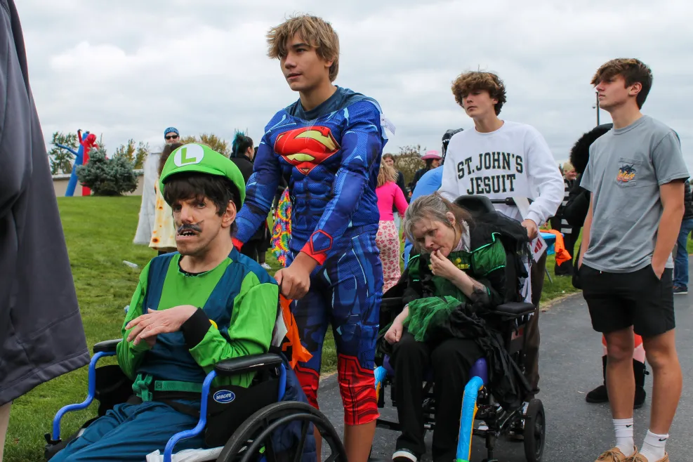 A group of men, including two in wheelchairs, dressed in Mario and Superman costumes, all smiling and enjoying a Halloween party. The group is gathered together, celebrating in a fun and inclusive atmosphere.