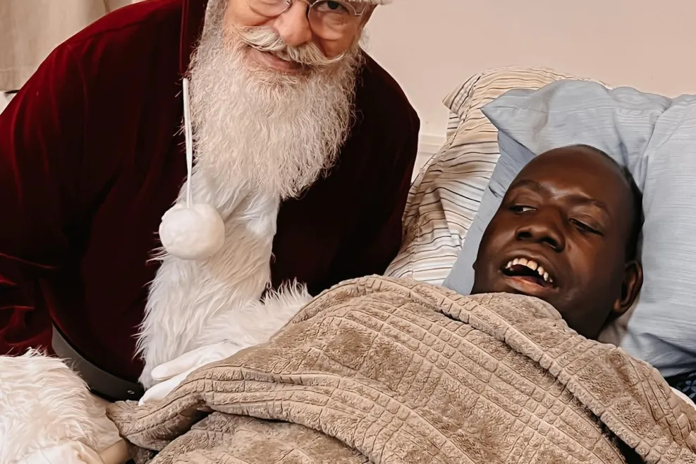 A smiling man wrapped in a cozy brown blanket lying on a bed, with a person dressed as Santa Claus leaning in beside him, smiling warmly
