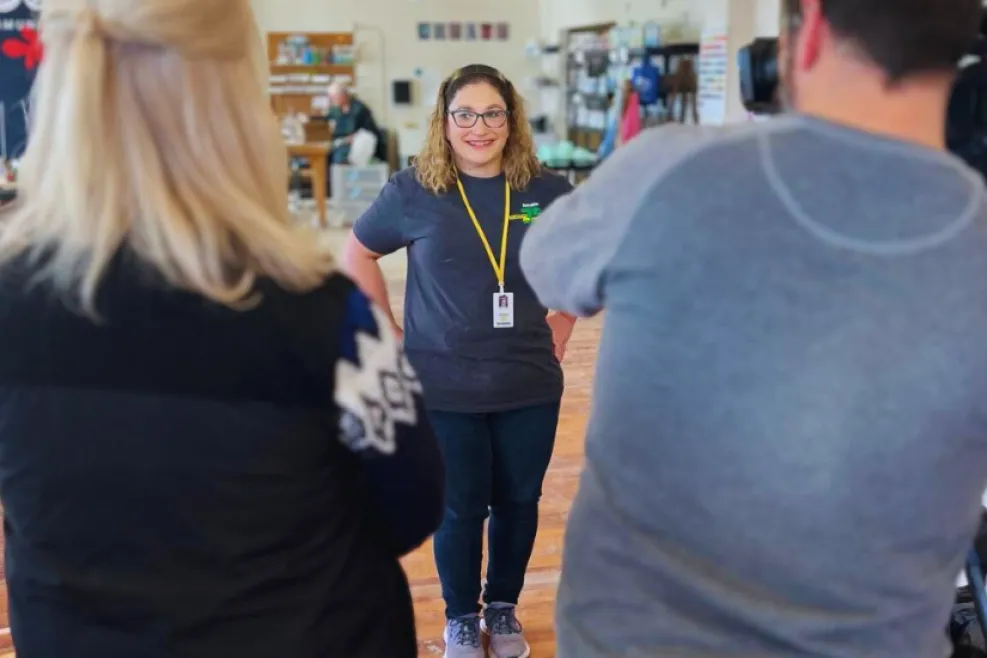 "A woman with curly hair, glasses, and a smile stands in the center of a well-lit workspace, wearing a gray t-shirt with a logo, a yellow lanyard, and an ID badge. She appears to be giving an interview, looking at a camera operated by a man in a gray shirt, while another person with blonde hair and a black vest stands nearby. The background features shelves with various items and an open layout with a wooden floor, suggesting a creative or retail environment."