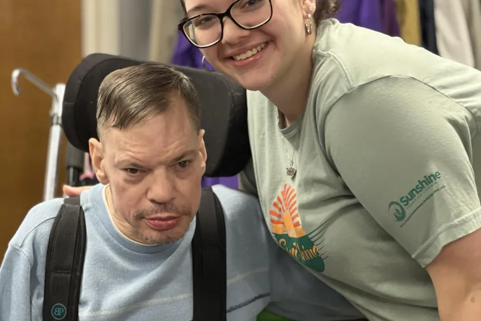 A young woman with dark hair in a bun, glasses, and a green Sunshine Communities t-shirt smiles as she leans next to a man in a wheelchair. The man, wearing a light blue sweater and black harness, holds a small red fabric item in his hands. Behind them, a clothing rack with various garments is visible in a well-lit indoor setting.
