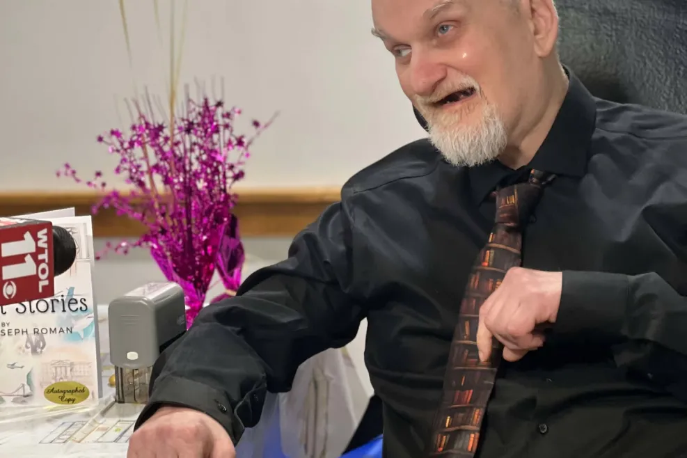 A man with a white beard and short hair sits at a decorated table, smiling and gesturing with his hand. He is dressed in a black shirt and a patterned tie featuring books. On the table beside him is a copy of 16 Short Stories by Jeffrey Joseph Roman, along with a stamp and festive decorations. Behind him, gold and white balloons float, adding to the celebratory atmosphere.