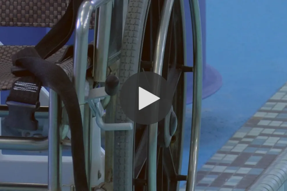 Close-up of a wheelchair next to a swimming pool, with a blue pool deck and mosaic tile edge visible in the background.