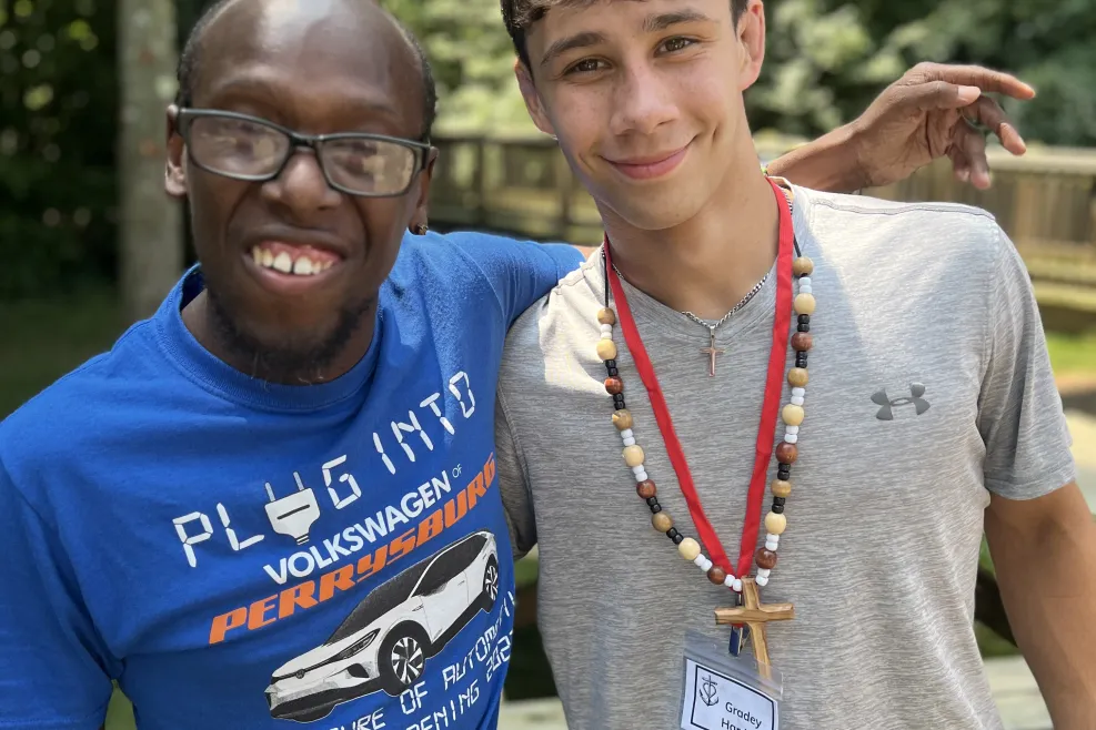 Alt text: Two young men smiling outdoors with arms around each other. The man on the left wears glasses and a blue “Plug Into Volkswagen Perrysburg” T-shirt, while the man on the right wears a gray Under Armour shirt, red lanyard, and wooden cross necklace. Trees and a wooden fence are blurred in the background.     