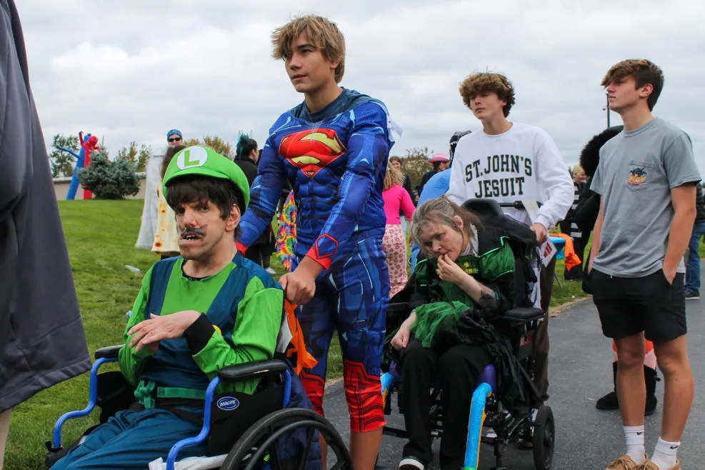 A group of people participate in an outdoor Halloween-themed event. Two individuals in wheelchairs are in the foreground.
