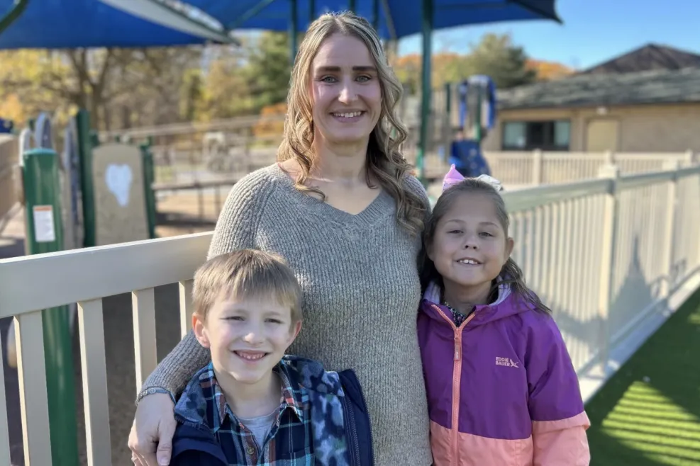 A woman stands outdoors at a playground with two children, one on each side of her.