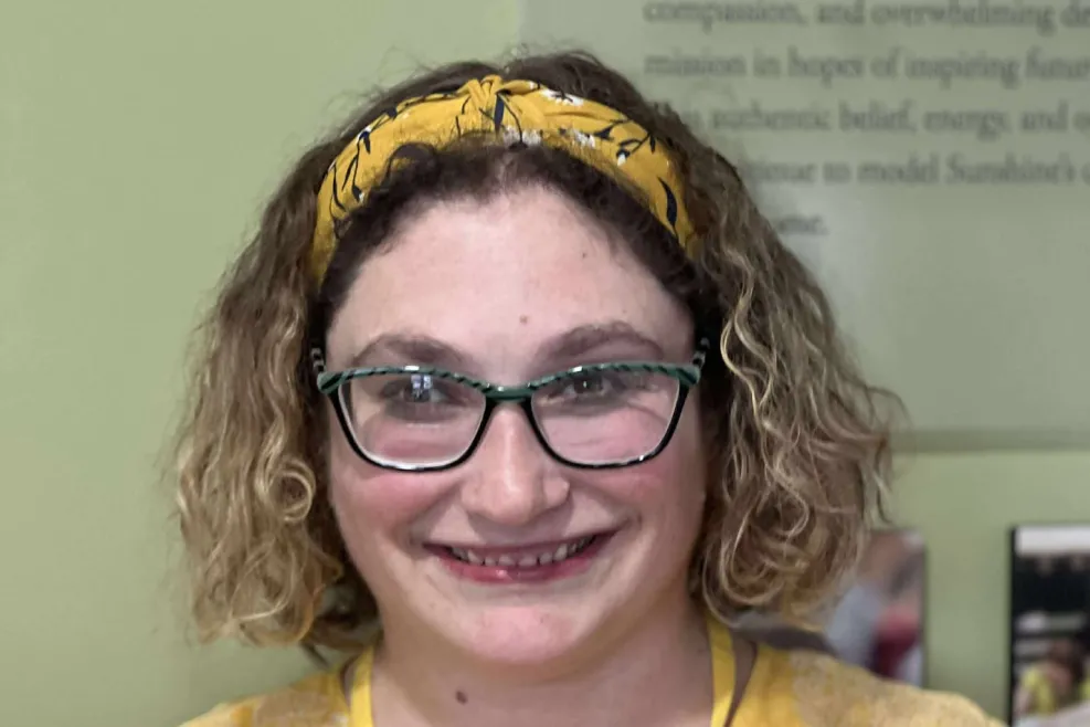 Melissa Jender smiles in a yellow top and matching headband, wearing glasses and a lanyard, in an indoor setting at Sunshine Communities.