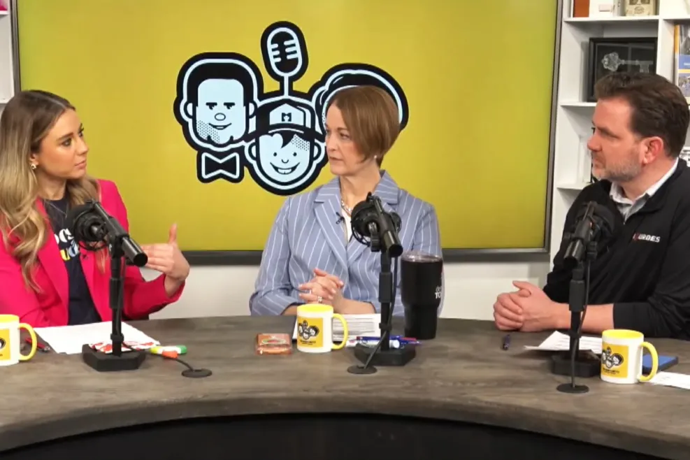 Three guests sit at microphones during a recording of The Four Hundred & Nineteen in a WGTE studio in Toledo, Ohio. A bright yellow screen with the show’s logo fills the background as the group talks around a curved table with branded mugs and notes.