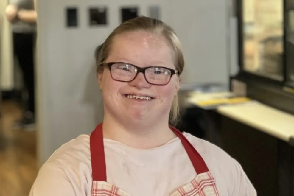 Person wearing glasses and a red plaid apron standing behind the counter at Georgette’s Café.