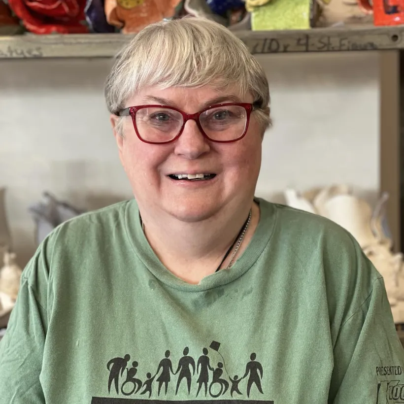 "Smiling woman with short gray hair and red glasses wearing a green Sunshine Communities 'Walk, Run & Roll' t-shirt, standing in front of colorful ceramic art pieces at Sunshine Studios in Maumee, Ohio."