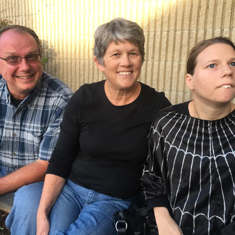 Smiling family sitting together outdoors — a man in a plaid shirt, a woman in a black top, and a young woman in a black spiderweb-patterned outfit, sitting against a brick wall.