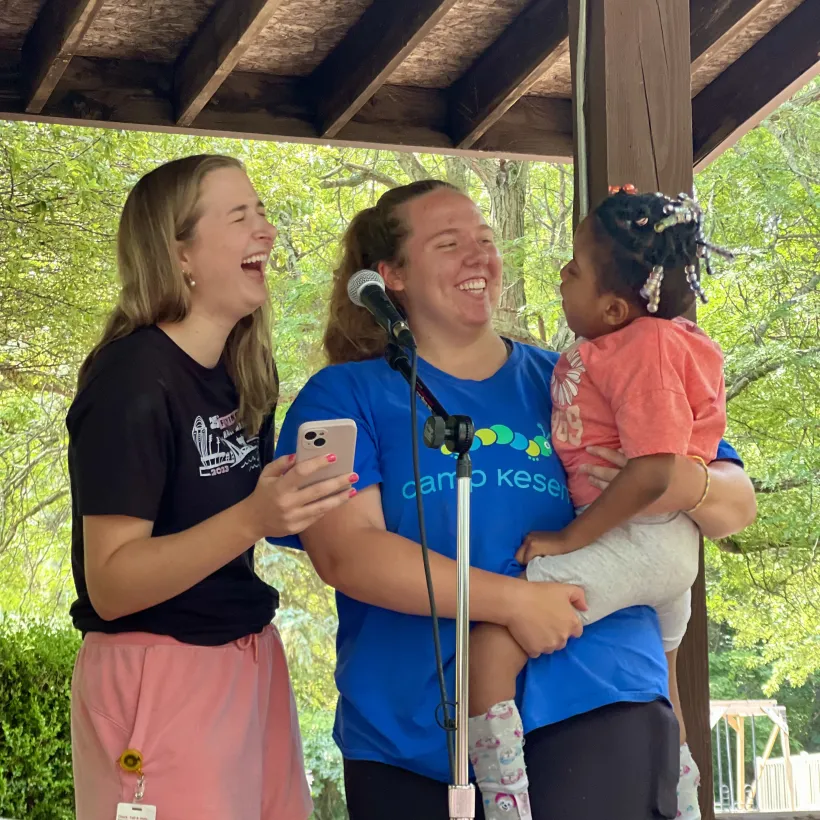 Two young women laughing together on stage at an outdoor camp event, one holding a toddler with beaded braids, near a microphone under a wooden pavilion.