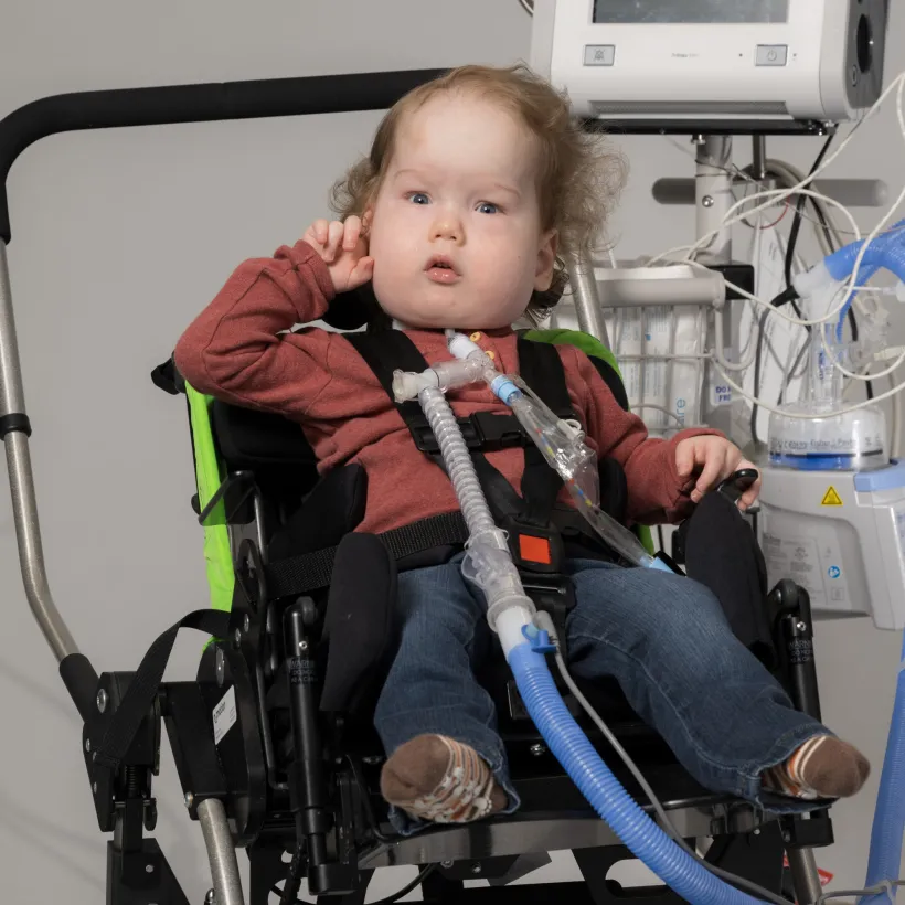 Young child with curly hair sitting in a specialized wheelchair, connected to a ventilator and medical tubing, wearing a maroon shirt, jeans, and brown socks.