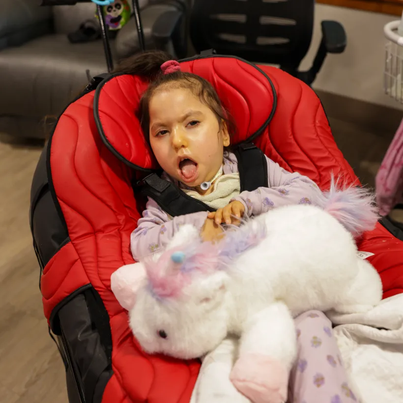 A young child lies comfortably in a padded red medical chair, supported with a harness. The child has a tracheostomy tube and rests with eyes partially open, tongue slightly out.