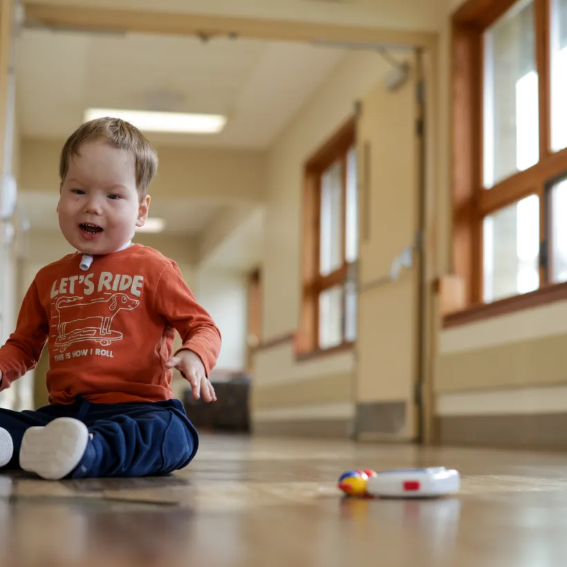 A young child with a tracheostomy sits on the floor of a bright hallway, smiling and playing with a small toy nearby.